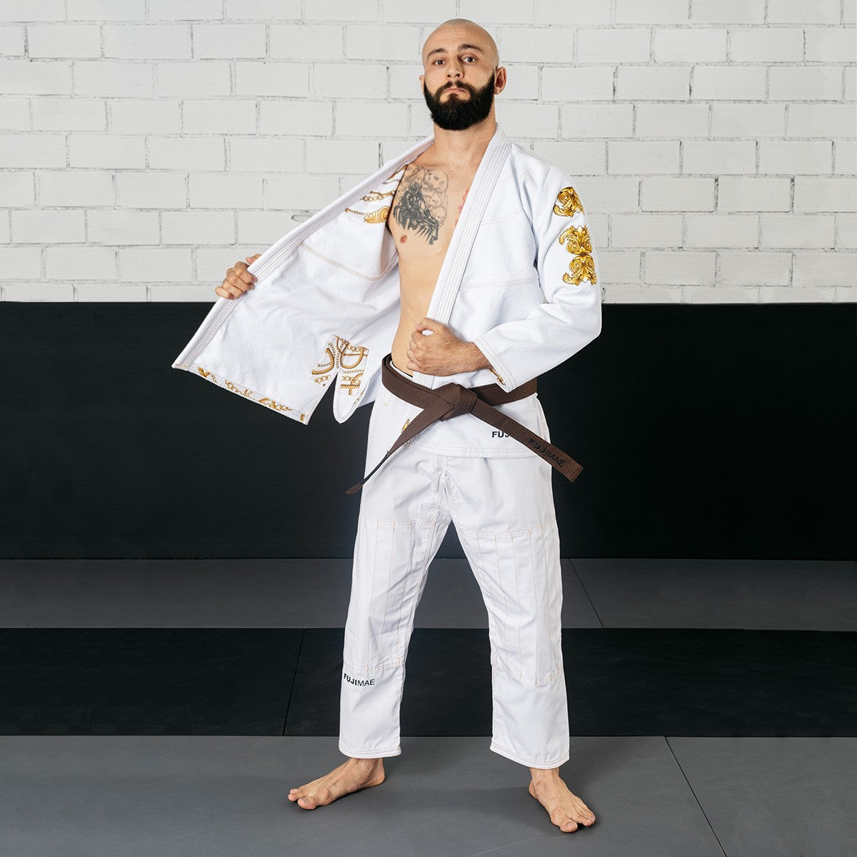 Man wearing white Shaka 21 BJJ Gi QS with gold embroidery and brown belt standing on mat against brick wall background
