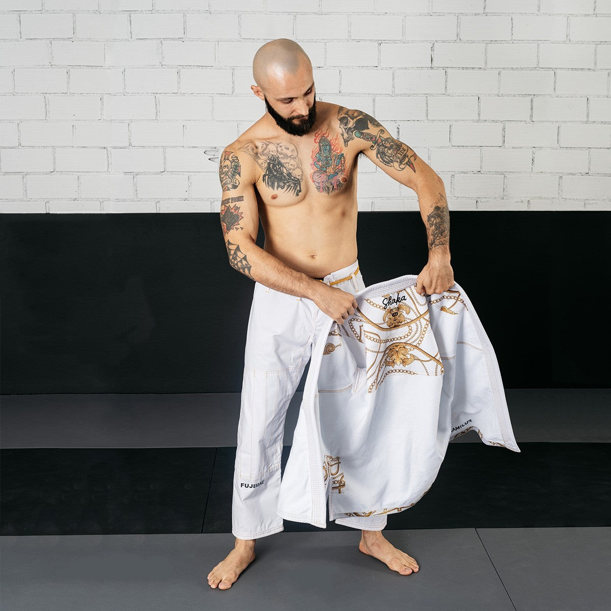 Man holding the white Shaka 21 BJJ Gi QS jacket with gold embroidery, wearing white BJJ pants with "FUJIMAE" logo, preparing to put on the gi in a martial arts training setting.