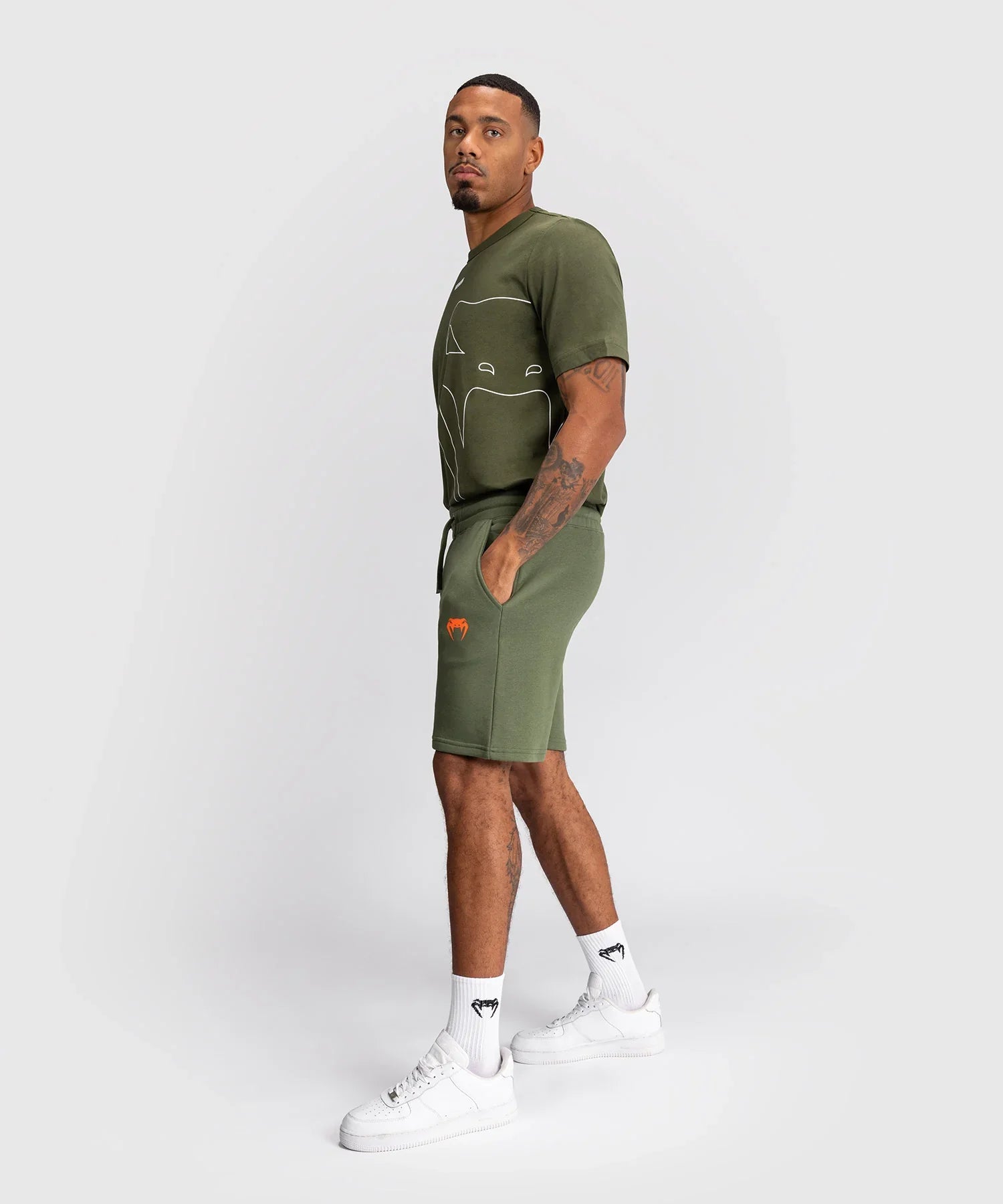 Man wearing khaki Shorts Venum Classic with elastic waistband, side and back pockets, and orange logo, paired with matching green t-shirt and white sneakers.