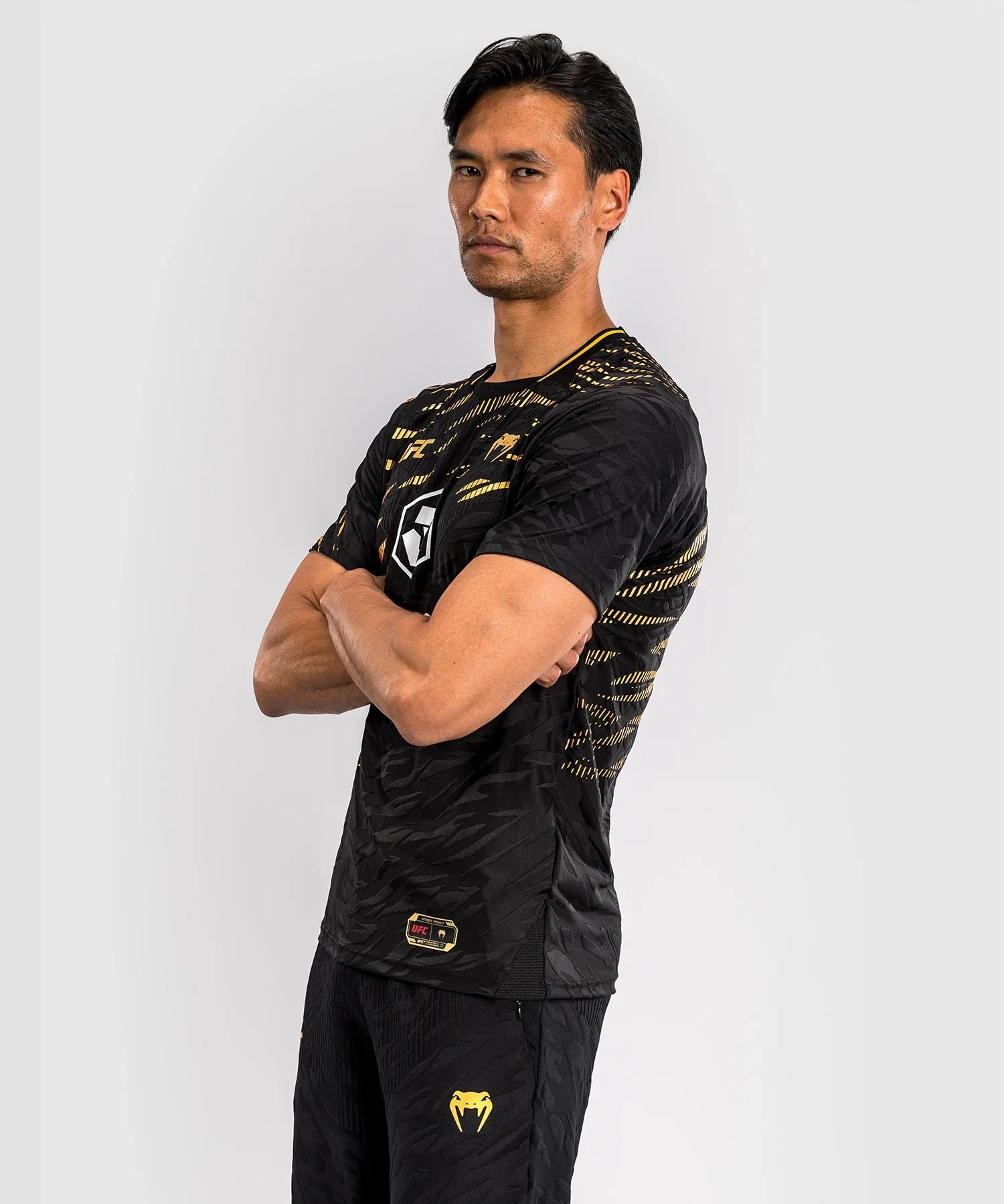 Man wearing black and gold UFC Fusion par Venum Dry-Tech T-shirt with short sleeves and logo print, standing with arms crossed against a plain background.