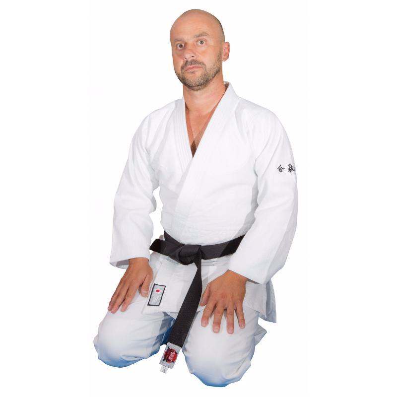 Man wearing white Kimono Aïkido-gi Dojo Master with black belt kneeling, showing short sleeves and embroidered Japanese Aïkido detail on left arm.